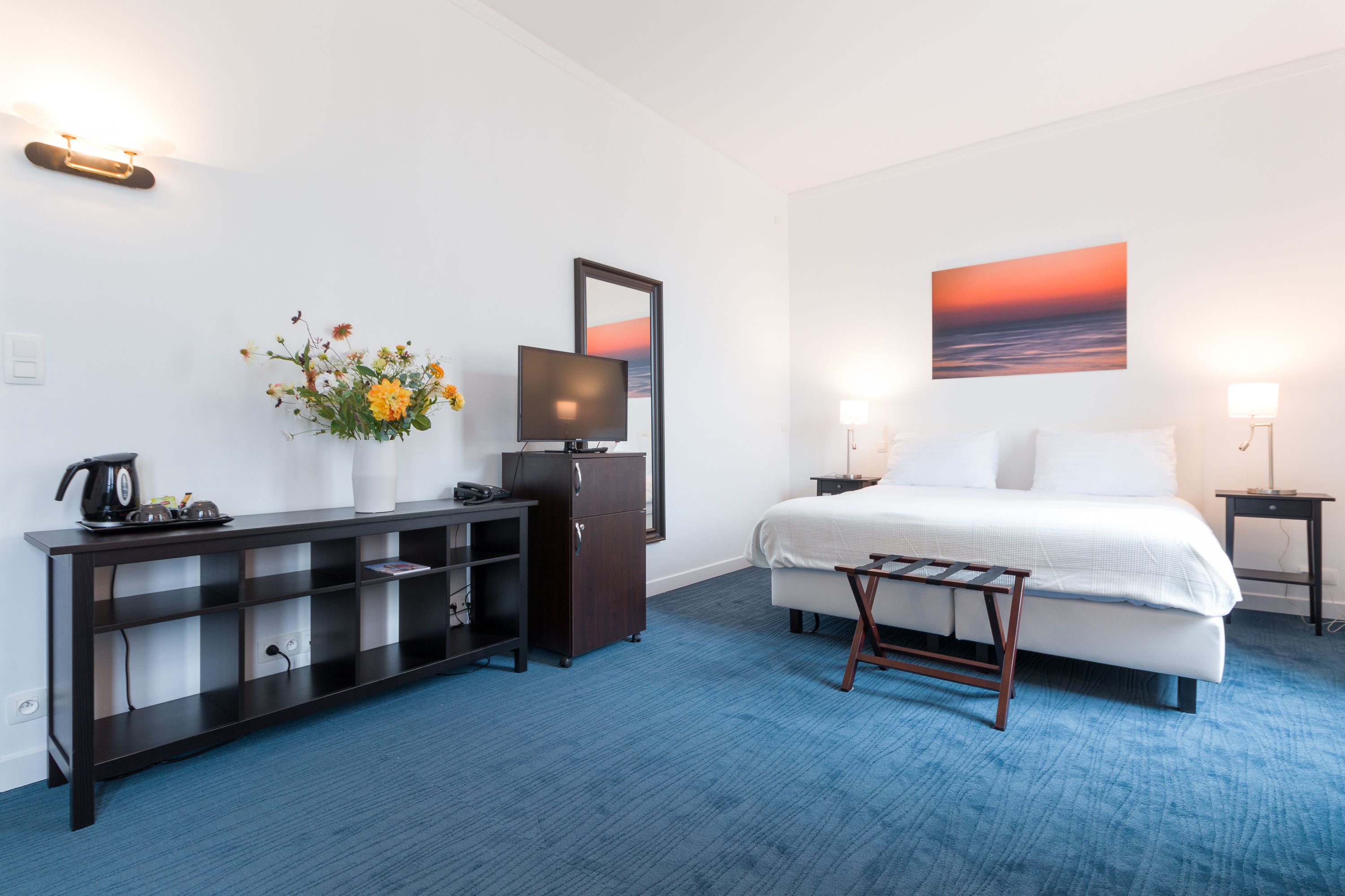 A hotel room with a bed, a TV, a desk with a computer, a coffee maker, a small table, and a wall-mounted shelf.