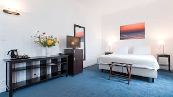 A hotel room with a bed, a TV, a desk with a computer, a coffee maker, a small table, and a wall-mounted shelf.