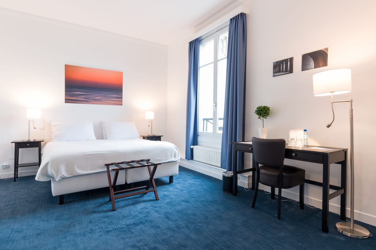 A hotel room with a bed, a desk, a chair, a lamp, a plant, and a window with curtains.