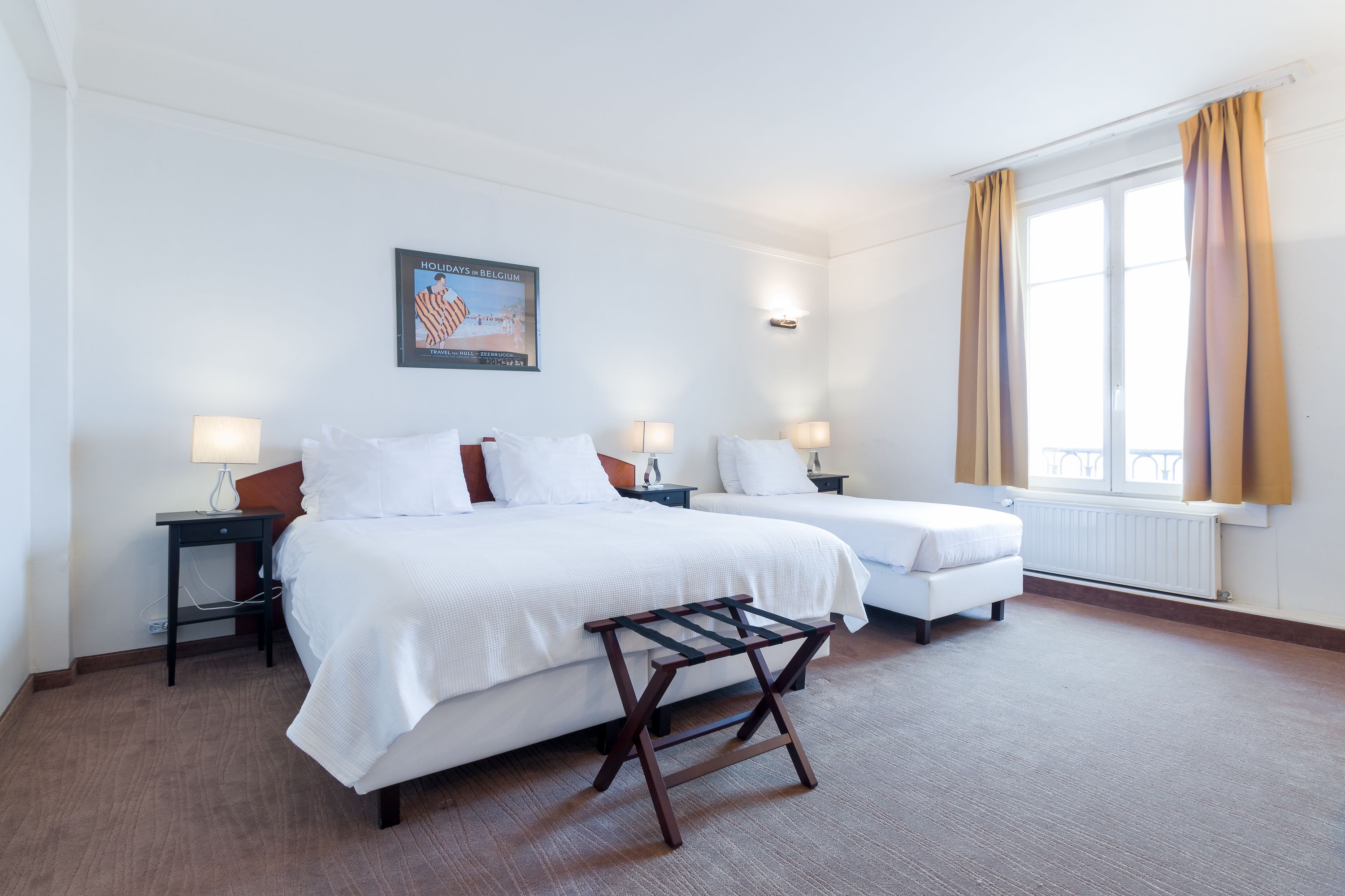 A hotel room with two beds, a wooden bench, a poster on the wall, and a window with curtains.