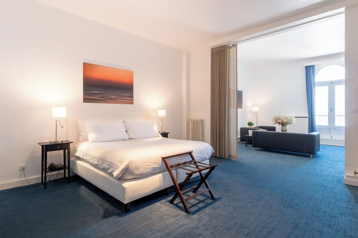 A hotel room with a large bed, two bedside lamps, a nightstand, a wooden chair, a sofa, and a view of the outdoors through a window.
