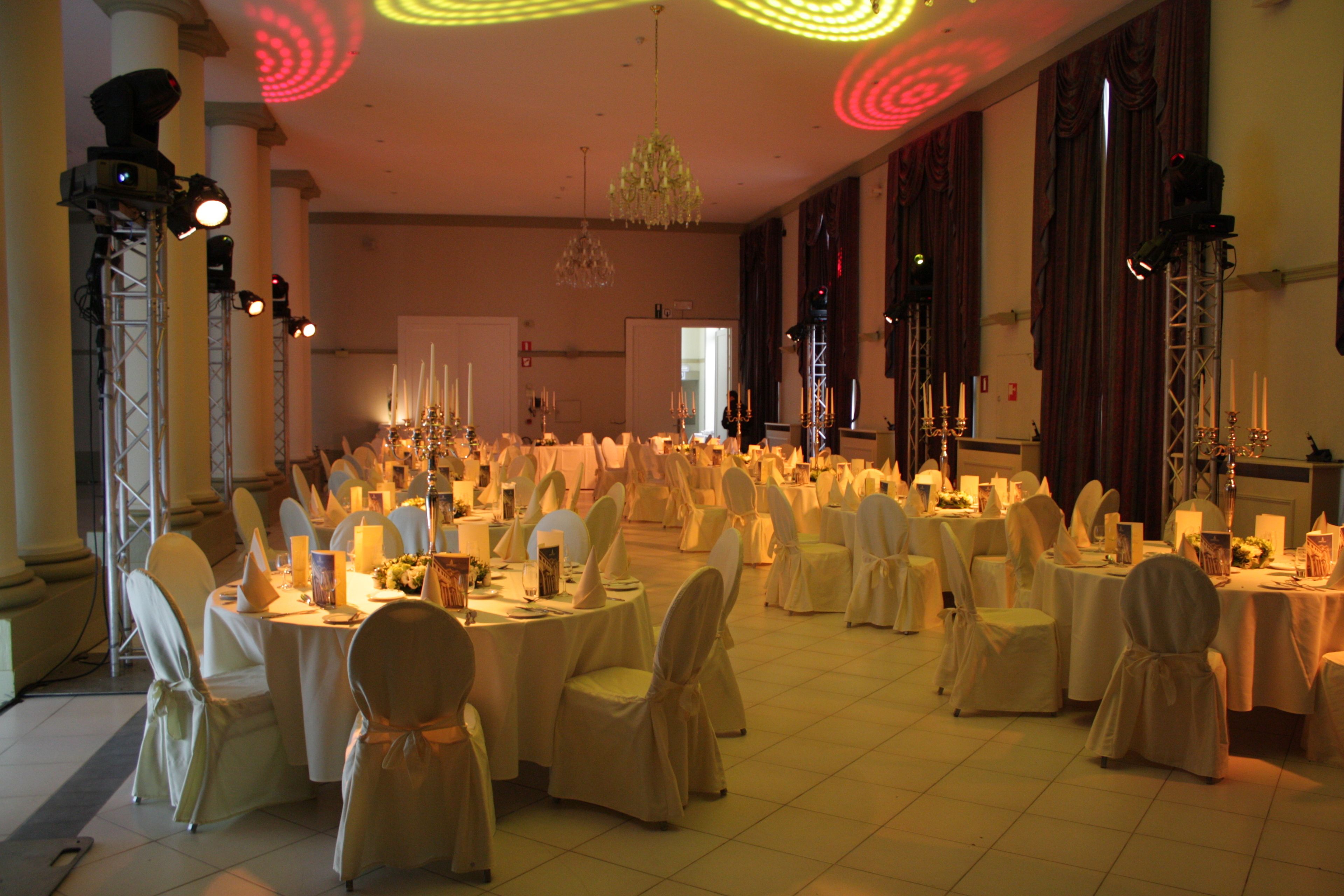 Una sala da banchetto con tavoli rotondi apparecchiati per un evento formale, con lampadari e luci da palcoscenico.