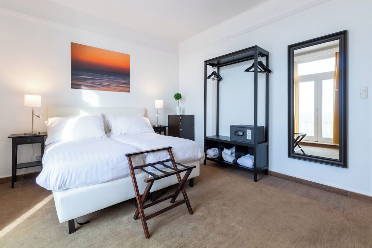 A modern hotel room with a large bed, bedside tables, a nightstand, a dresser, a mirror, and a view of the sunset.