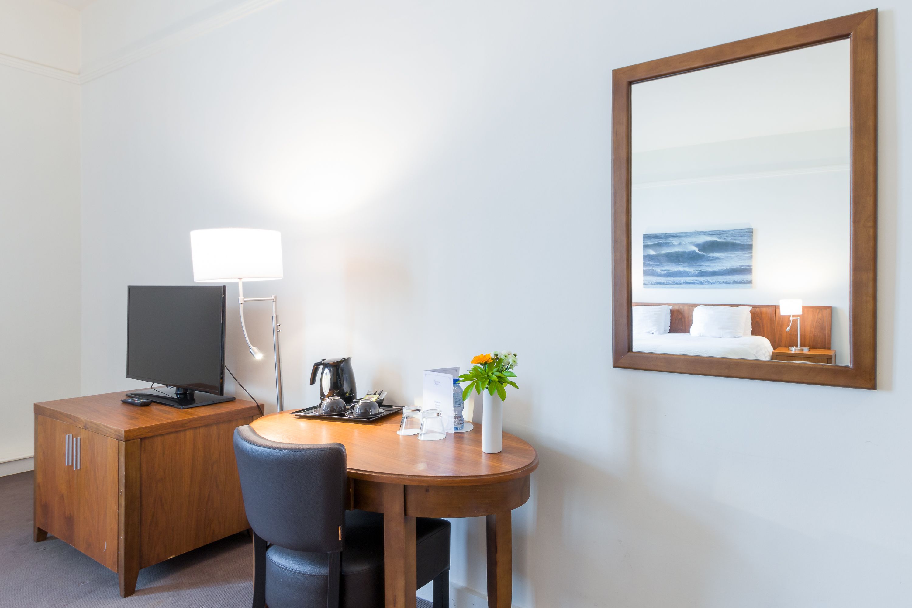 A hotel room with a wooden desk, a television, a coffee maker, a vase with flowers, and a large mirror reflecting a bed and a nightstand.