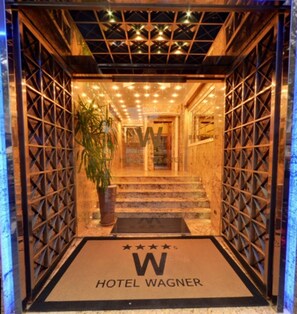 L’entrée d’un hôtel, avec une façade ornée d’une grille métallique, un pot de fleurs et un paillasson portant le nom de l’hôtel.