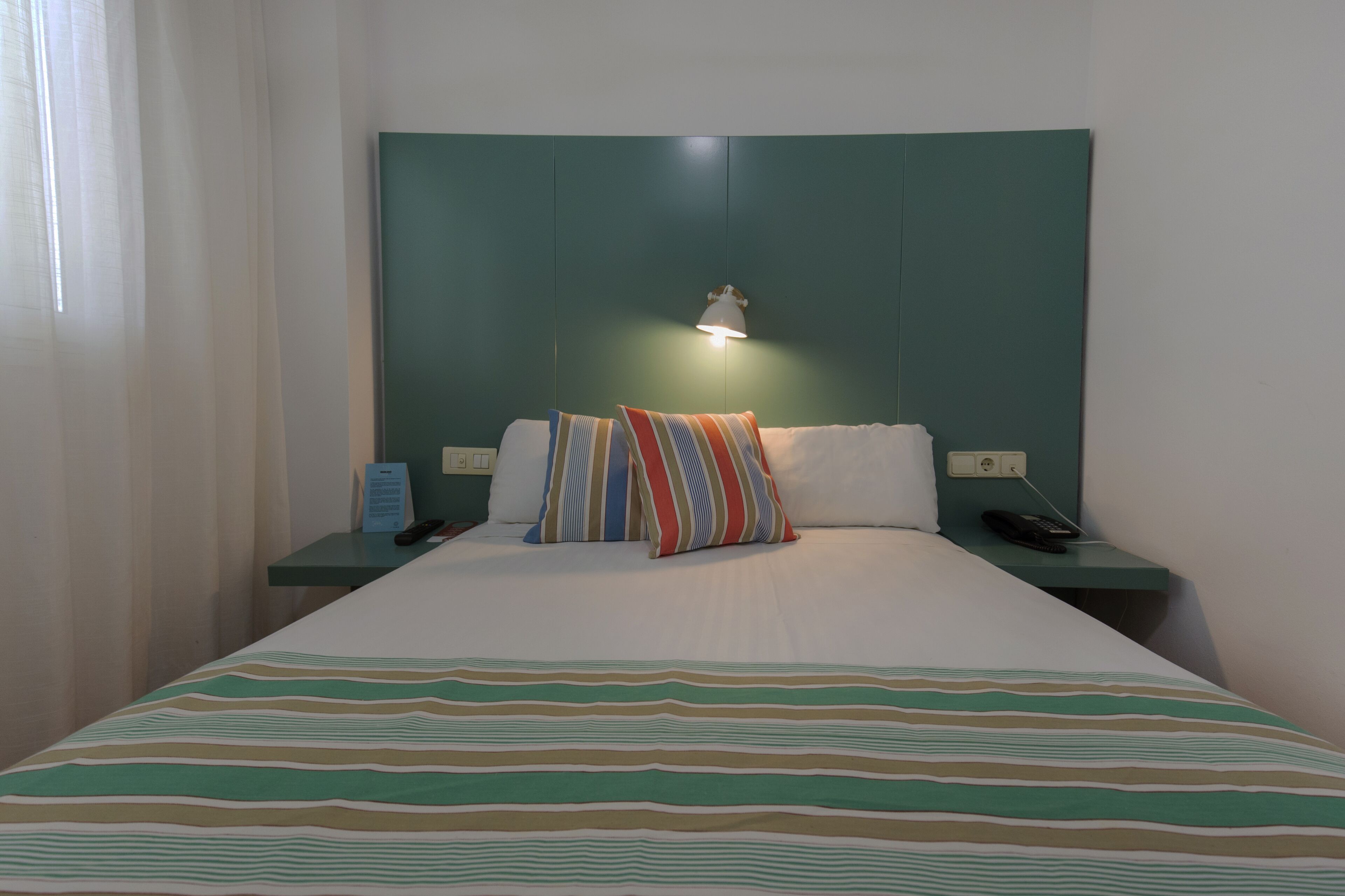 Una habitación de hotel con una cama, ropa de cama a rayas, un cabecero verde azulado, una lámpara de noche, un teléfono y una mesita de noche.
