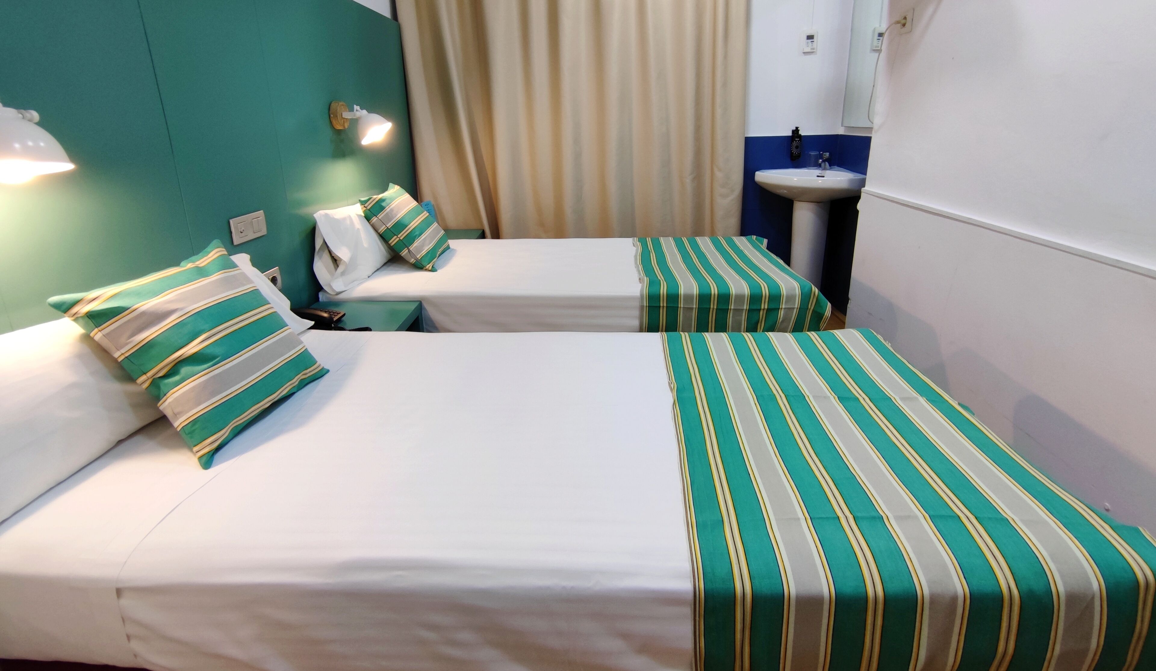 A hotel room with two beds, each with striped bedding, a bedside lamp, a small table, and a bathroom with a sink and mirror.