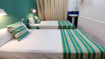 A hotel room with two beds, each with striped bedding, a bedside lamp, a small table, and a bathroom with a sink and mirror.