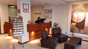 Zona de recepción de un hotel con un mostrador de madera, una pantalla digital, un área de asientos con sillones de cuero y un cuadro abstracto grande en la pared.