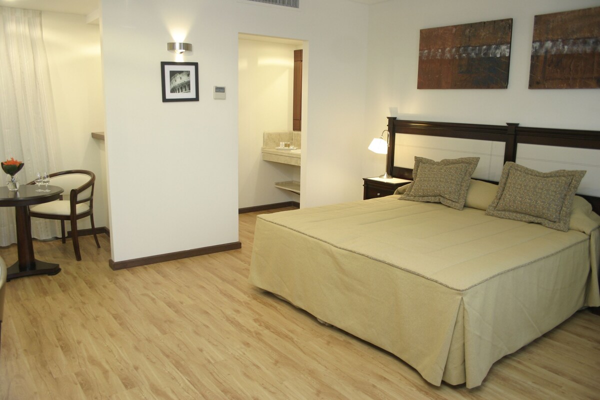 A hotel room with a bed, a desk with a chair, a nightstand, a wall-mounted lamp, and a framed picture.