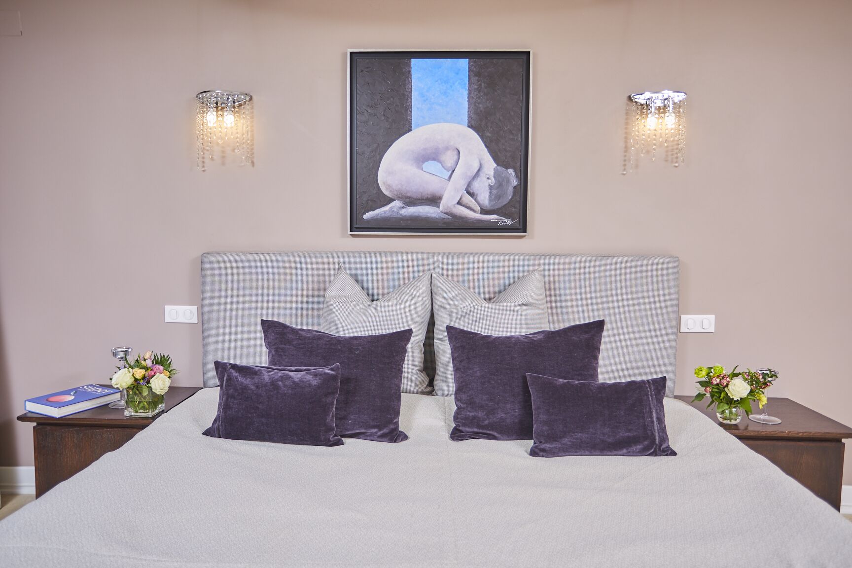 Un lit bien fait, avec une tête de lit à carreaux, deux oreillers violets et une œuvre d’art encadrée au-dessus.