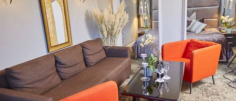 A hotel room with a brown sofa, orange armchair, and a small table with wine glasses. There is a mirror on the wall and a window with a view of the city.
