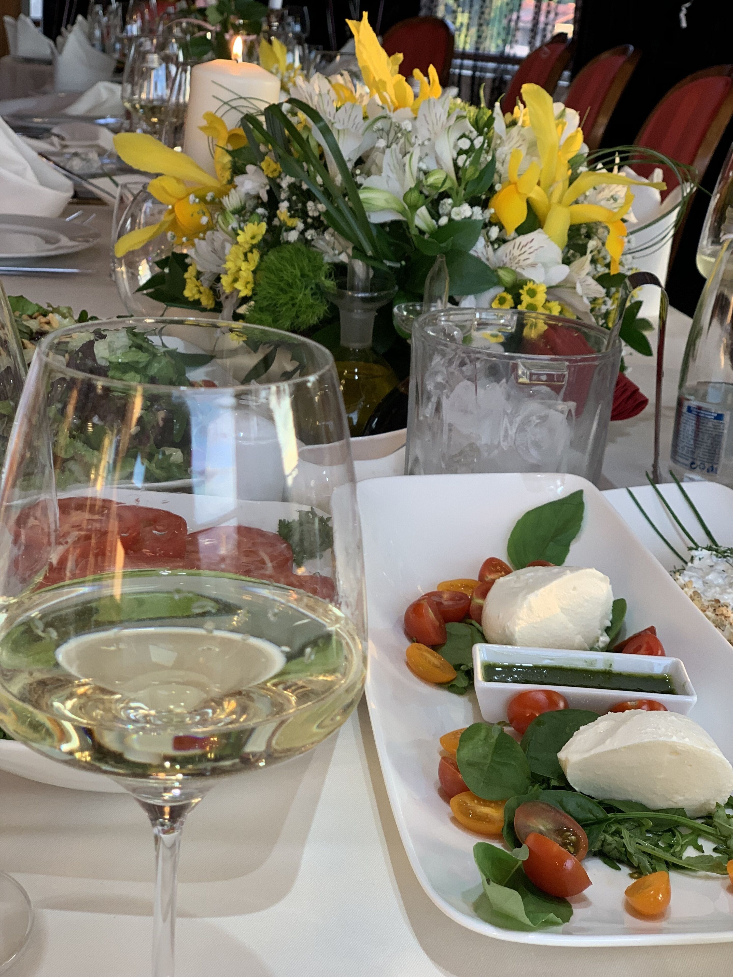 Een eettafel gedekt met glazen servies, een bord met tomaten en mozzarella, en een vaas met bloemen.