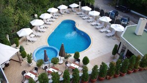 Piscina cercada por espreguiçadeiras e guarda-sóis, com plantas em vasos e área de estar com piso de azulejos.