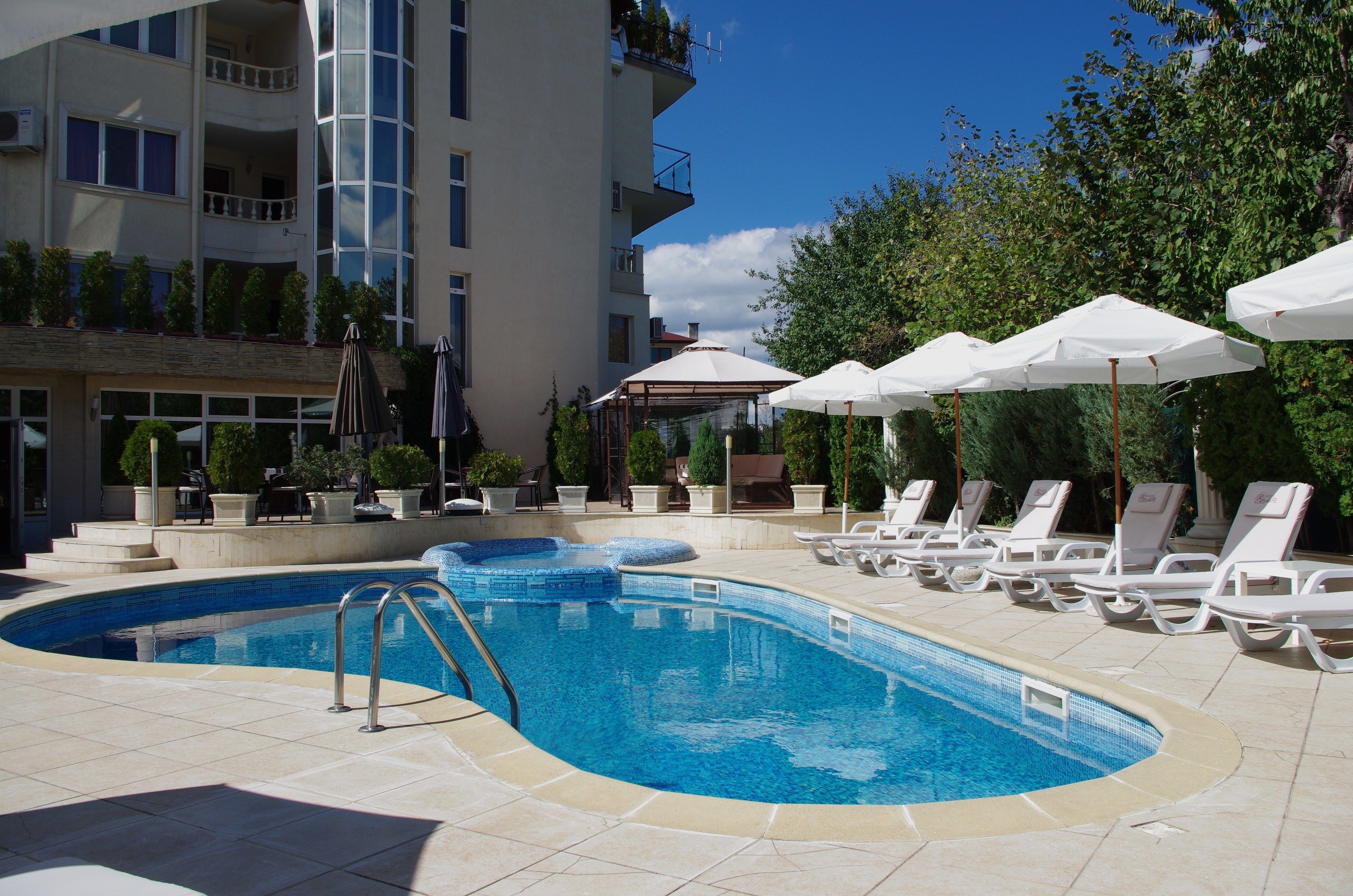 Une piscine entourée de chaises longues et de parasols, elle-même bordée d’un bâtiment doté de balcons et de plantes en pot.