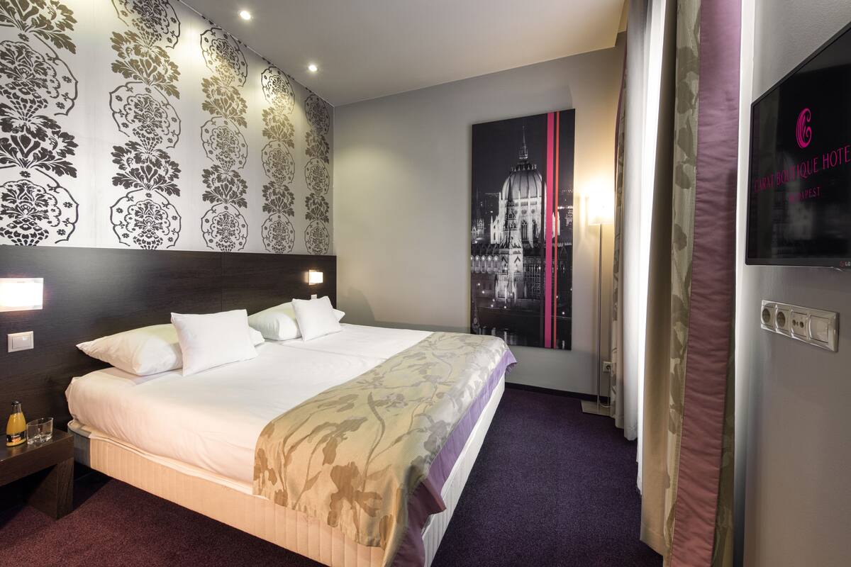 Une chambre d’hôtel avec un grand lit, une table de chevet avec une bouteille et un verre, un mur orné d’une affiche représentant un paysage urbain, et un téléviseur fixé au mur.