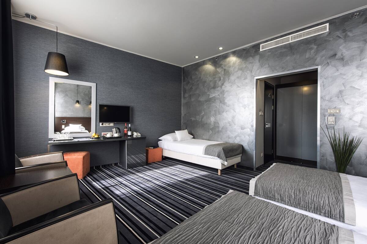 Une chambre d’hôtel moderne équipée d’un lit, d’un bureau avec un ordinateur, d’un miroir, d’une télévision et d’une grande fenêtre.