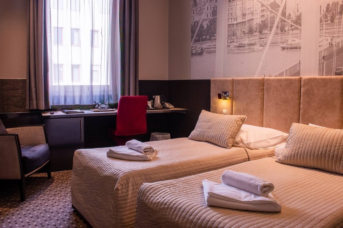 Une chambre d’hôtel avec deux lits, un bureau, une chaise et une grande fenêtre avec des rideaux.