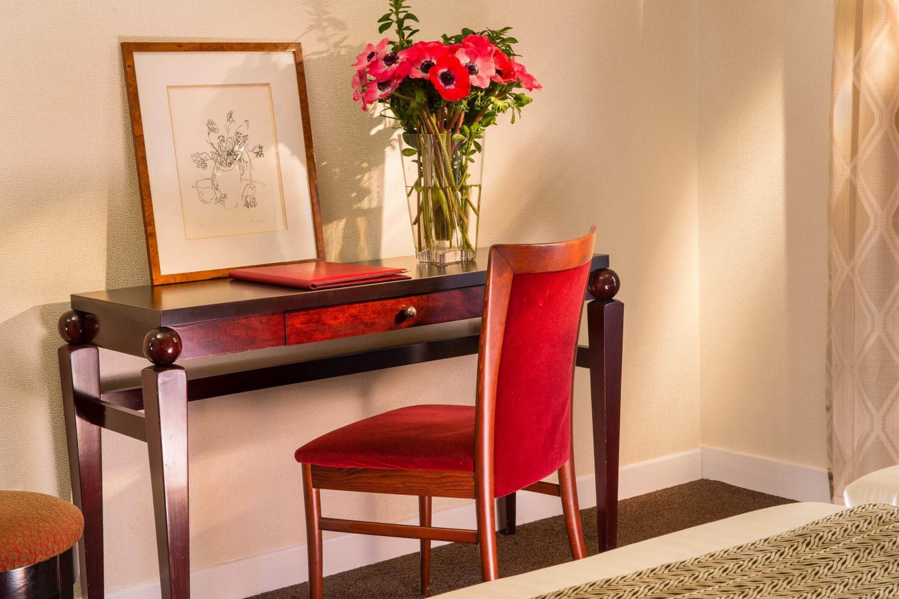 A desk with a framed drawing, a vase with red flowers, and a red chair.
