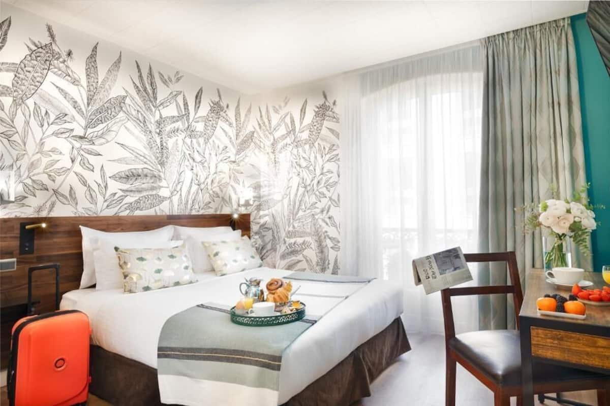 A hotel room with a large bed, a wooden headboard, a breakfast tray on the bed, a chair, a table with fruits, and a wall with a leaf pattern.
