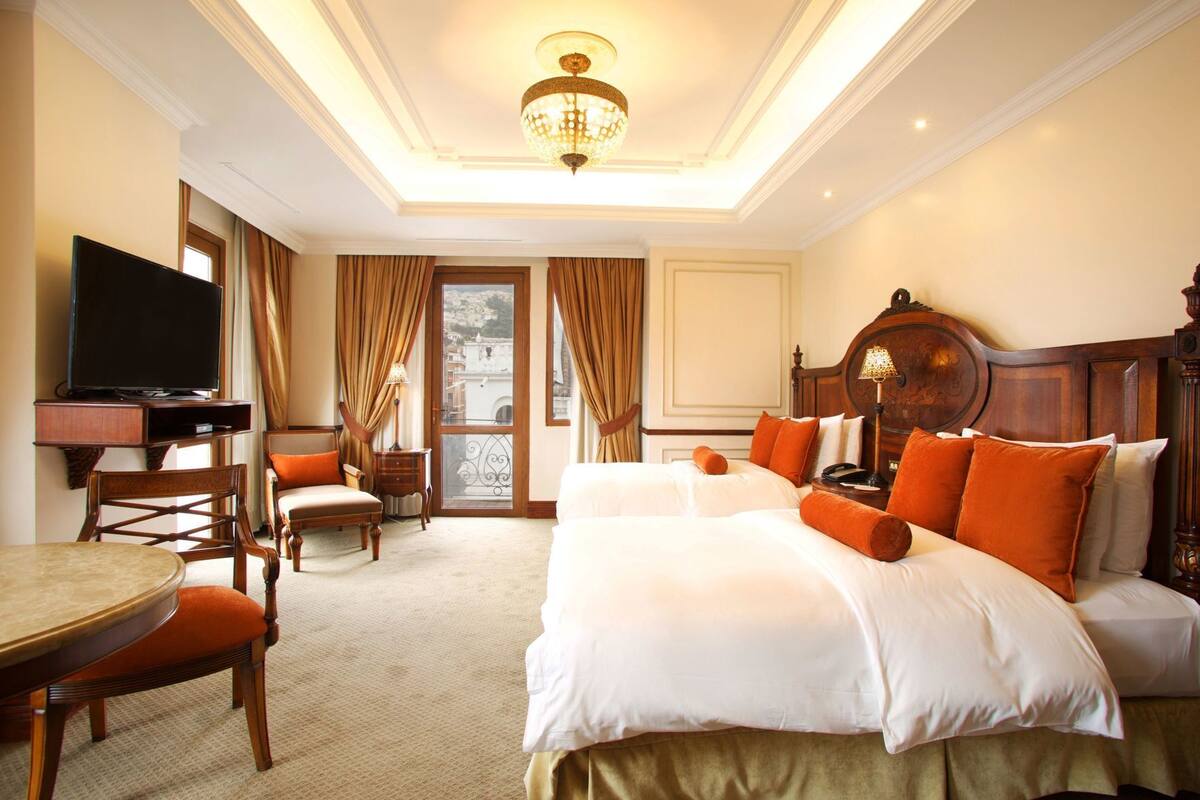 A hotel room with a large bed, a television, a dining table, and a view of a cityscape through the window.