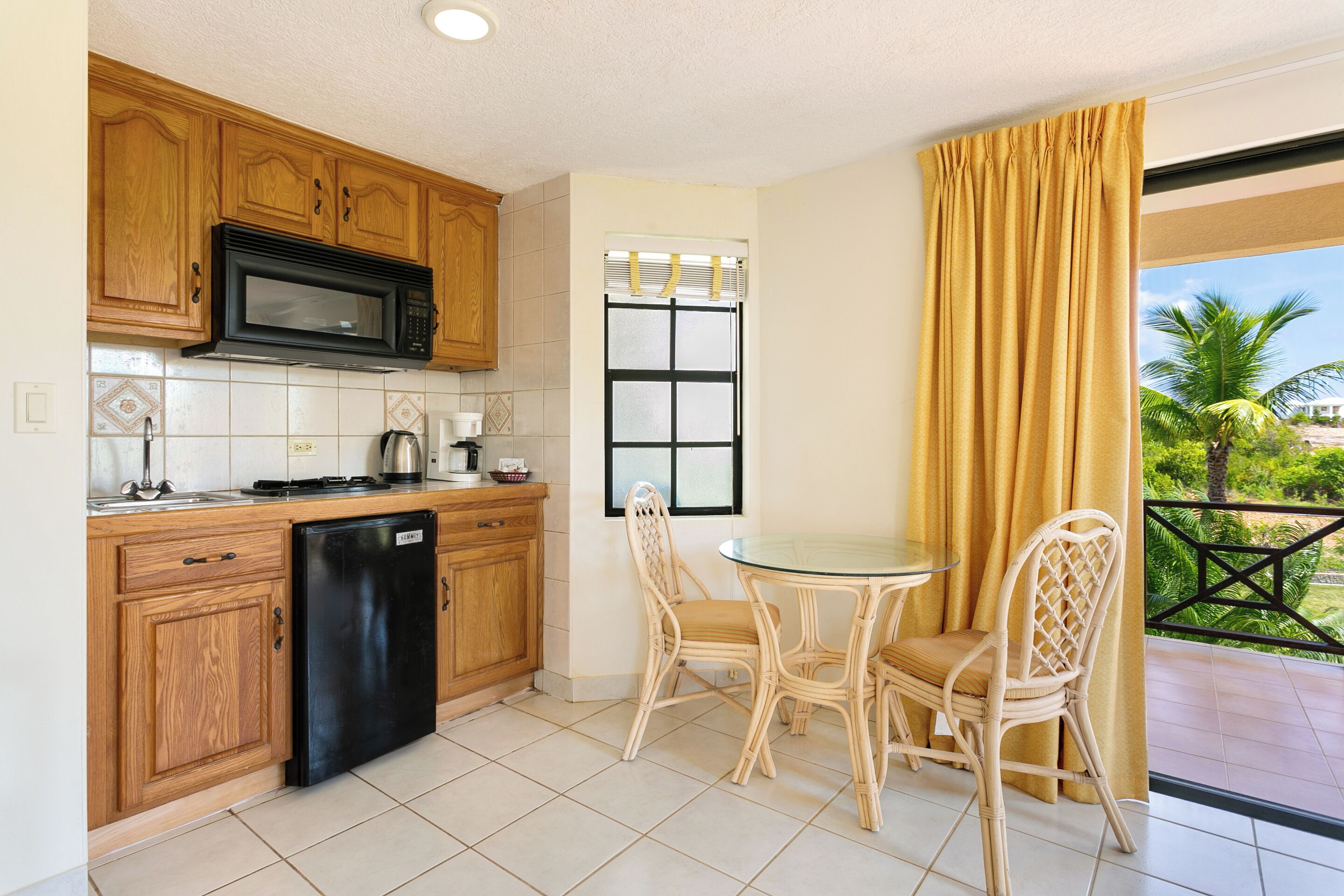 A kitchen with wooden cabinets, a black microwave, and a black refrigerator. There is a dining table with two chairs and a window with blinds.