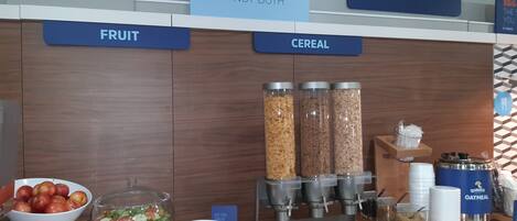 A breakfast bar with labeled sections for fruit and cereal, including bowls of apples and a salad, and dispensers for Quaker Oatmeal and other cereals.