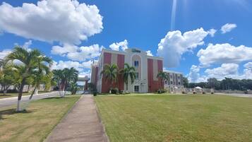 Un edificio grande con una entrada destacada, rodeado de un césped cuidado y palmeras, bajo un cielo azul despejado con algunas nubes.