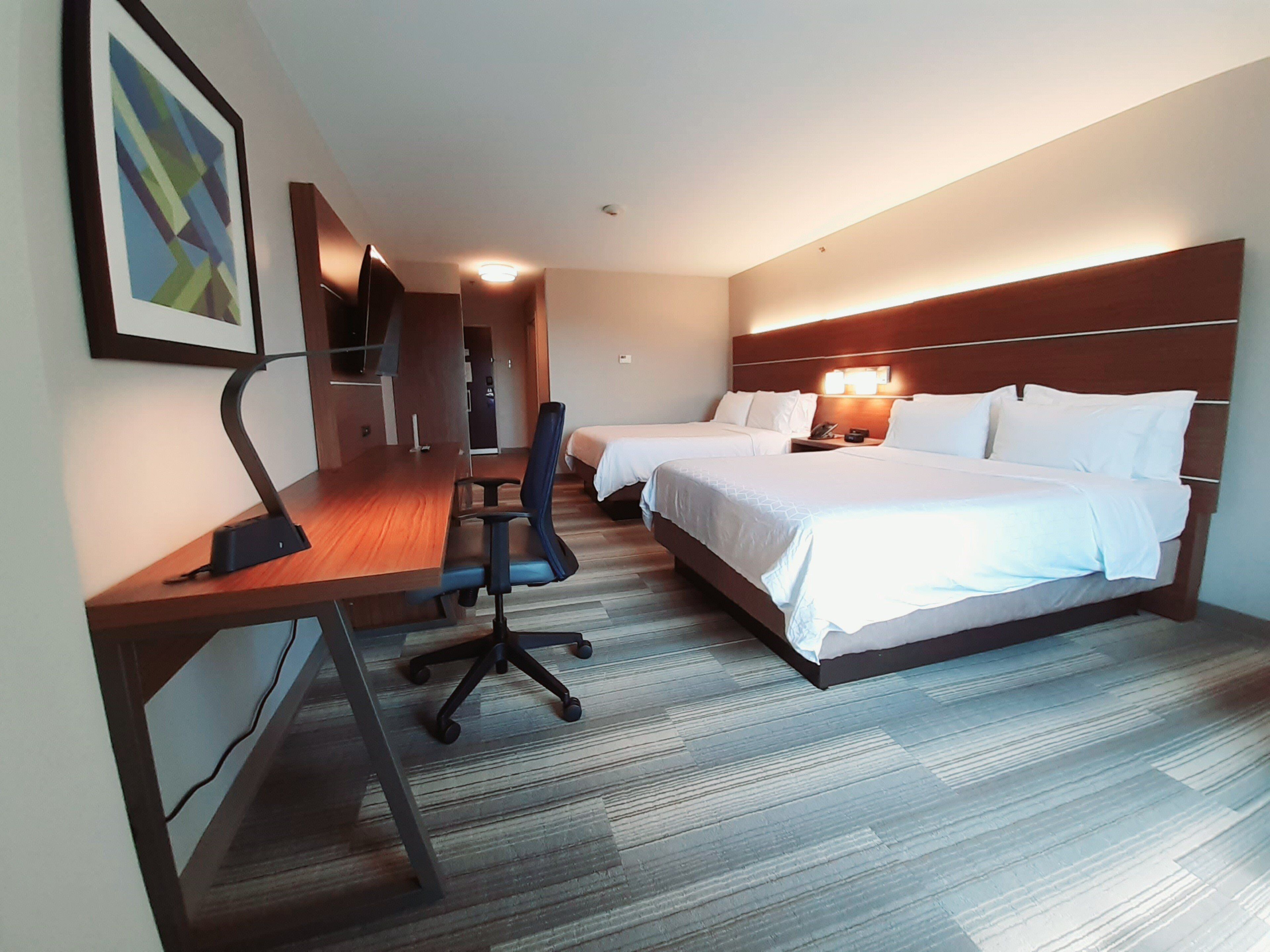 Habitación de hotel con un escritorio de madera, dos camas, una silla y una mesita de noche con lámpara.