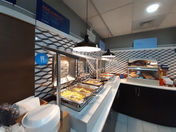 Zona de bufé con varias estaciones de comida, incluyendo una estación de gofres y una barra de ensaladas. Se observan carteles que indican opciones de autoservicio, y un letrero azul con el texto 'The Start You Need Express'.