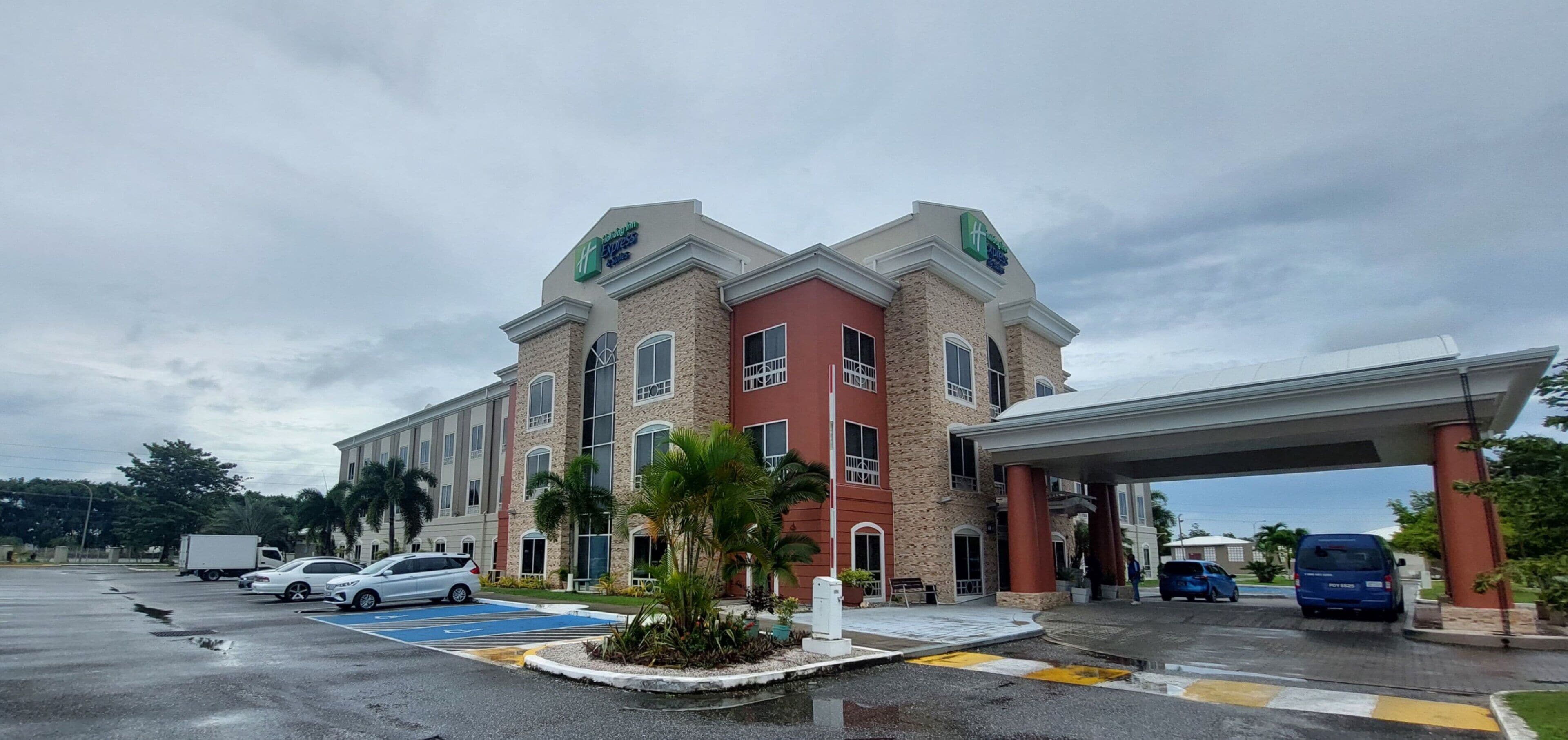 A hotel with a red and beige facade, a covered entrance, and a parking lot with several cars.