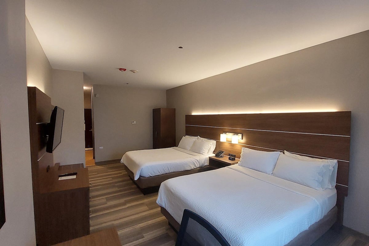 A hotel room with two beds, a desk with a chair, a TV, and a headboard with built-in lighting.