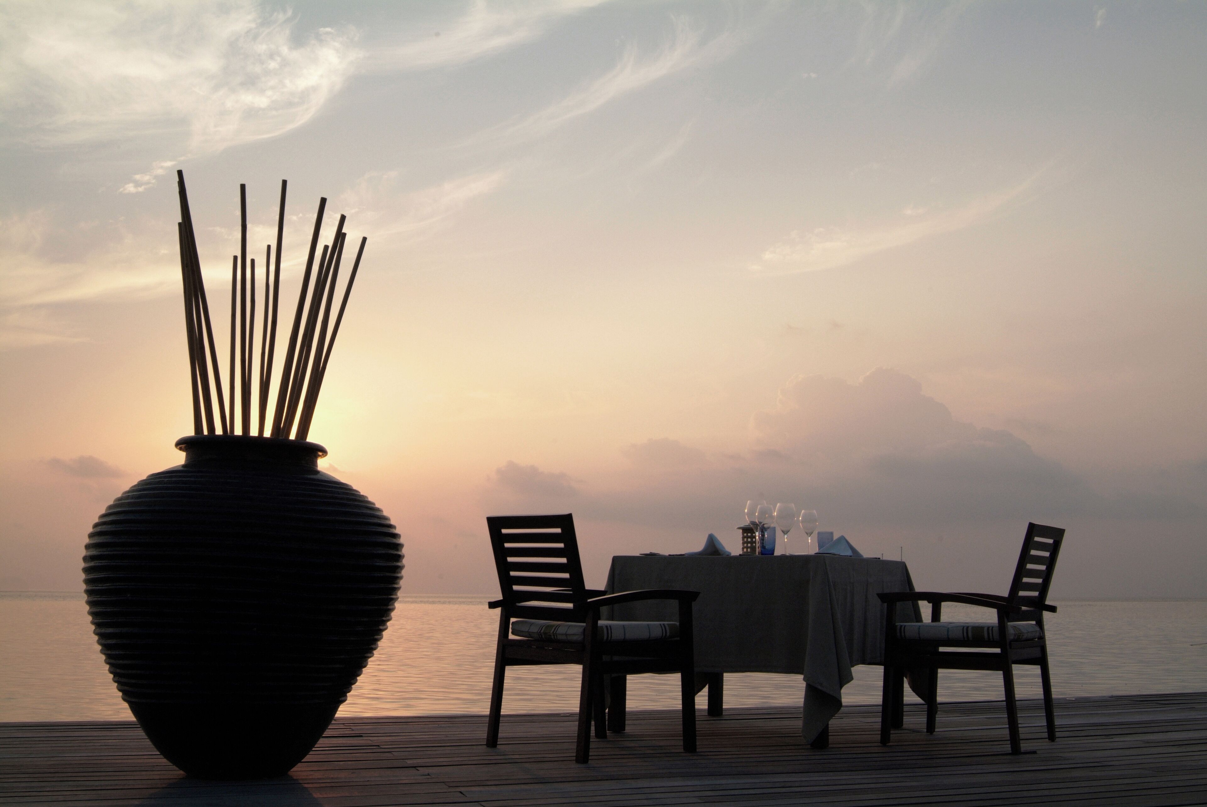 A dining table set for two with a large vase of sticks, overlooking a body of water.
