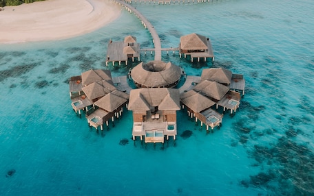 Vista aérea de bungalós sobre el agua con una piscina central, rodeados de aguas turquesas y una playa de arena.