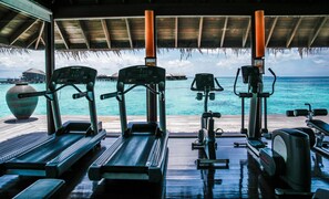 A gym with treadmills and exercise equipment, overlooking a turquoise sea and overwater bungalows. - Coco Bodu Hithi (Bodu Hithi)