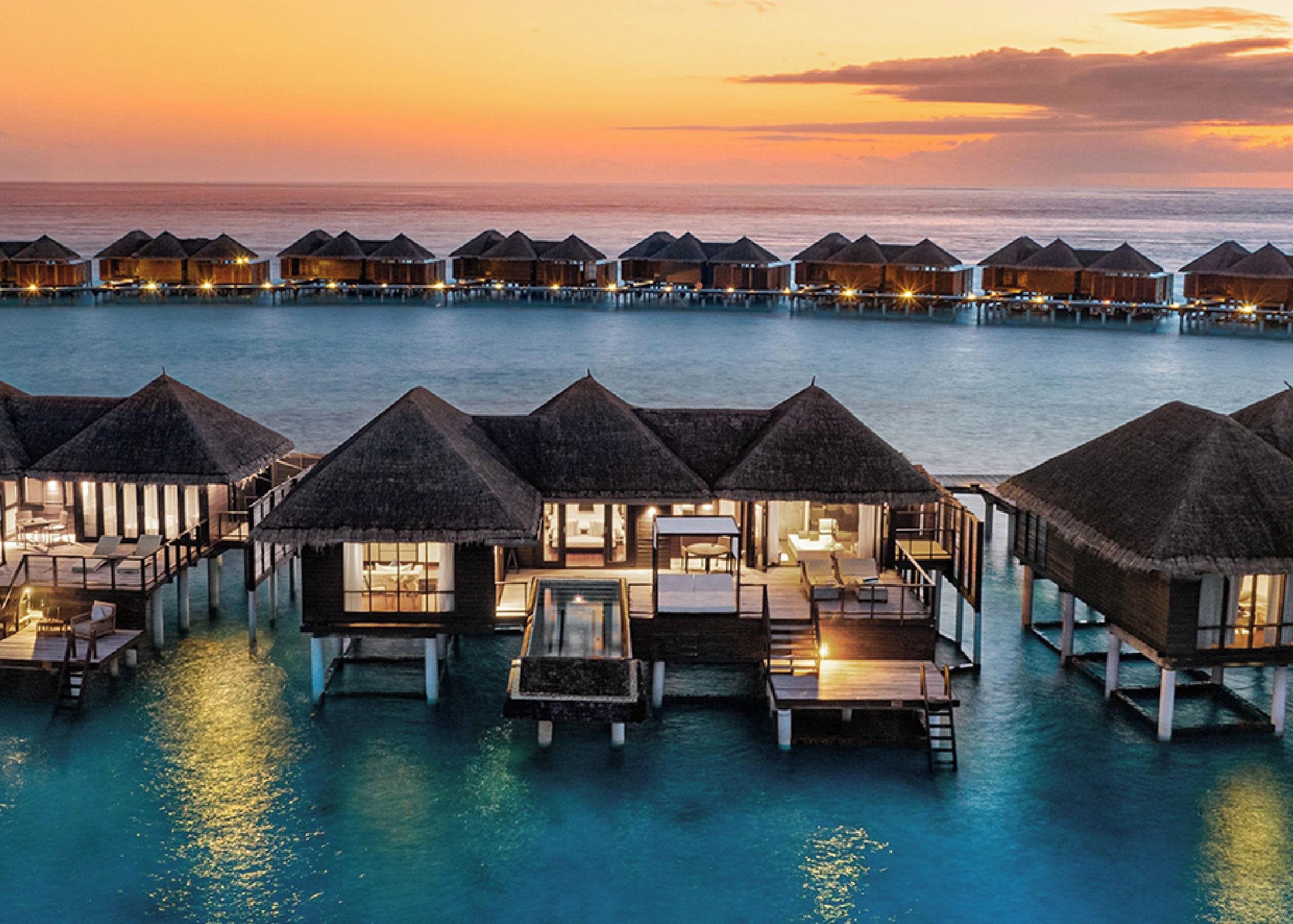 Wasserbungalows mit Reetdächern, verbunden durch Wege, mit Blick auf ein ruhiges Meer bei Sonnenuntergang.