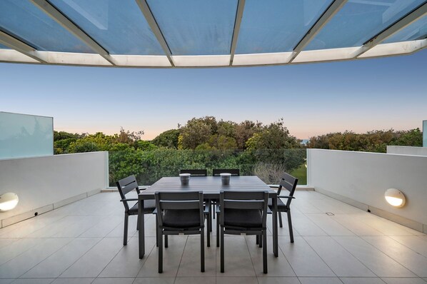 A modern outdoor patio with a table and chairs, surrounded by a glass enclosure, offering a view of a lush garden and trees. - Ramada By Wyndham Marcoola Beach (Marcoola)