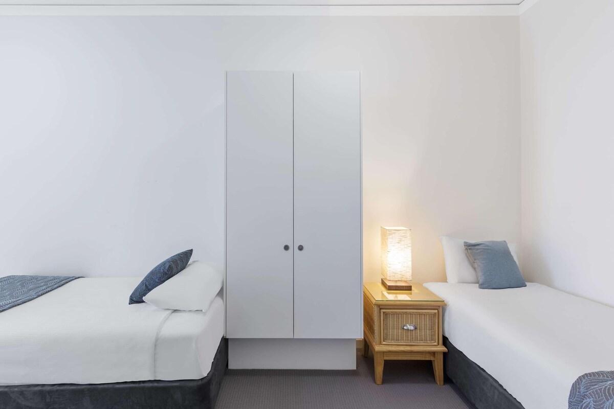 Habitación de hotel con dos camas individuales, un armario, una mesita de noche con lámpara y una pequeña mesita auxiliar de madera.