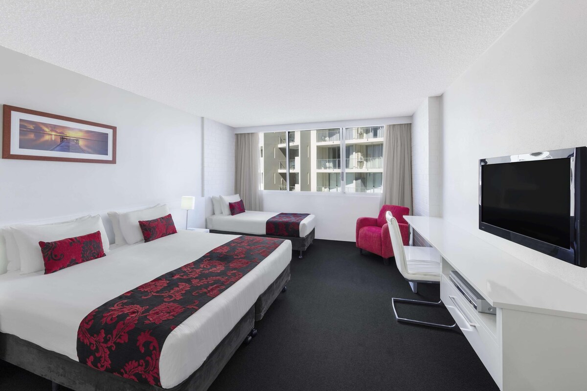 Habitación de hotel con dos camas, un escritorio, una silla, un televisor y una ventana con vista a edificios.