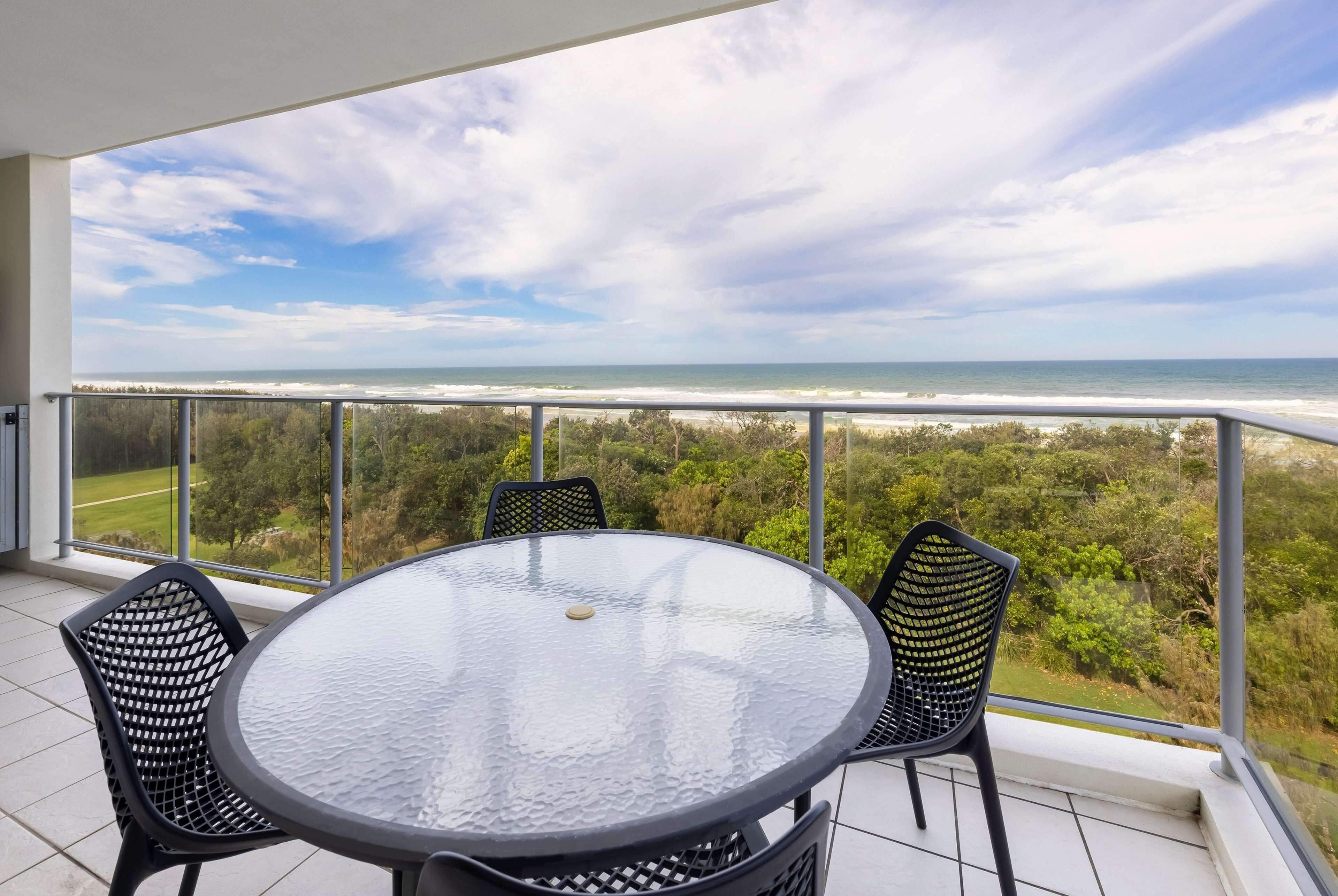 A balcony with a round glass table and four chairs, overlooking a beach and ocean.