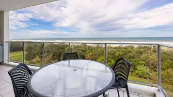 A balcony with a round glass table and four chairs, overlooking a beach and ocean.