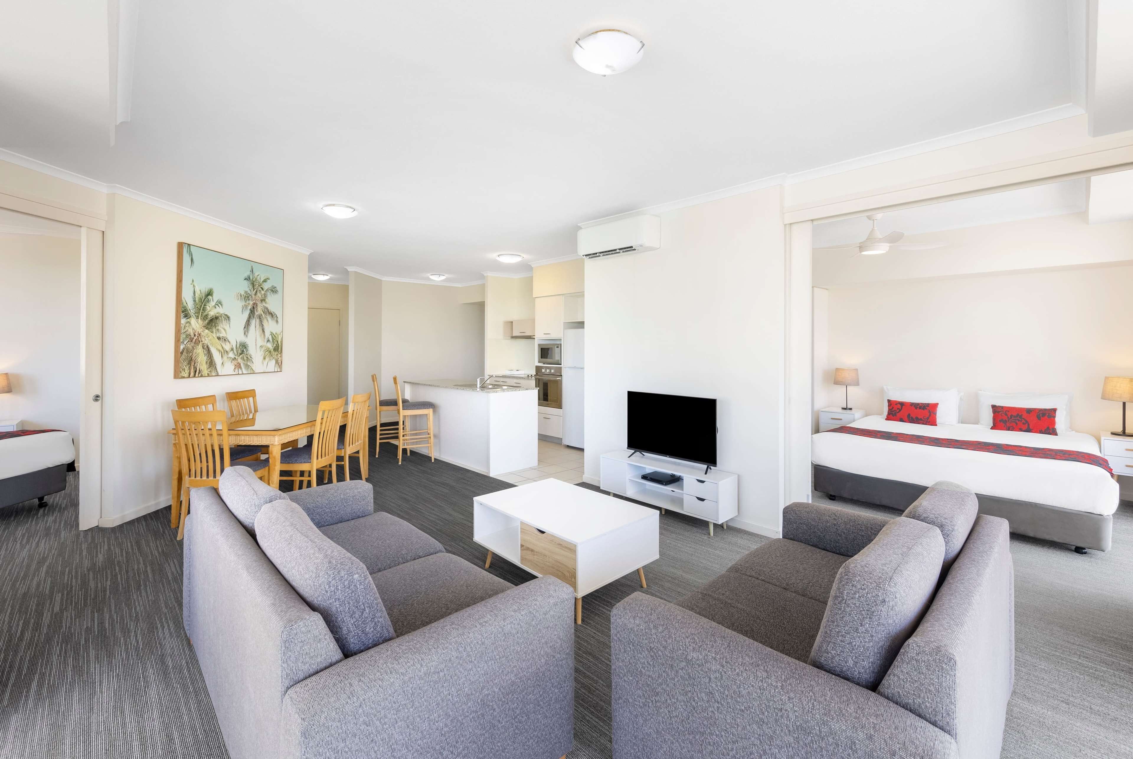 A modern hotel room with a sofa, TV, dining area, and a bed with red pillows.