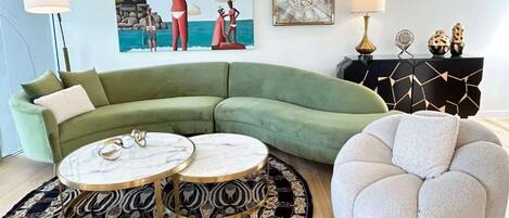 Una sala de estar moderna con un sofá seccional verde, dos mesas de centro redondas, un otoman blanco, un tapete estampado, un cuadro con una escena de playa y una mesita de noche de diseño geométrico.