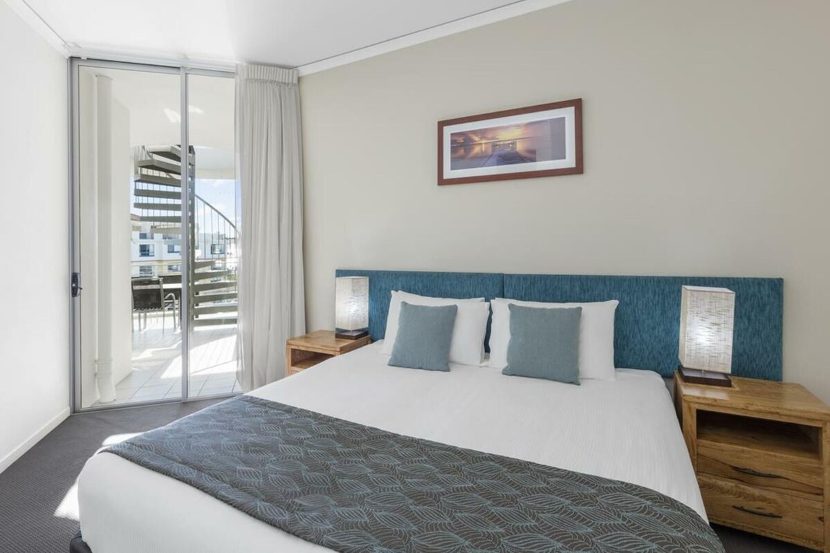 Habitación de hotel con una cama grande, dos mesitas de noche, vista a una escalera de caracol y un cuadro en la pared.