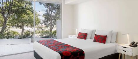 Una habitación de hotel moderna con una cama grande, ropa de cama blanca, una manta roja y negra estampada y una mesita de noche con una lámpara.
