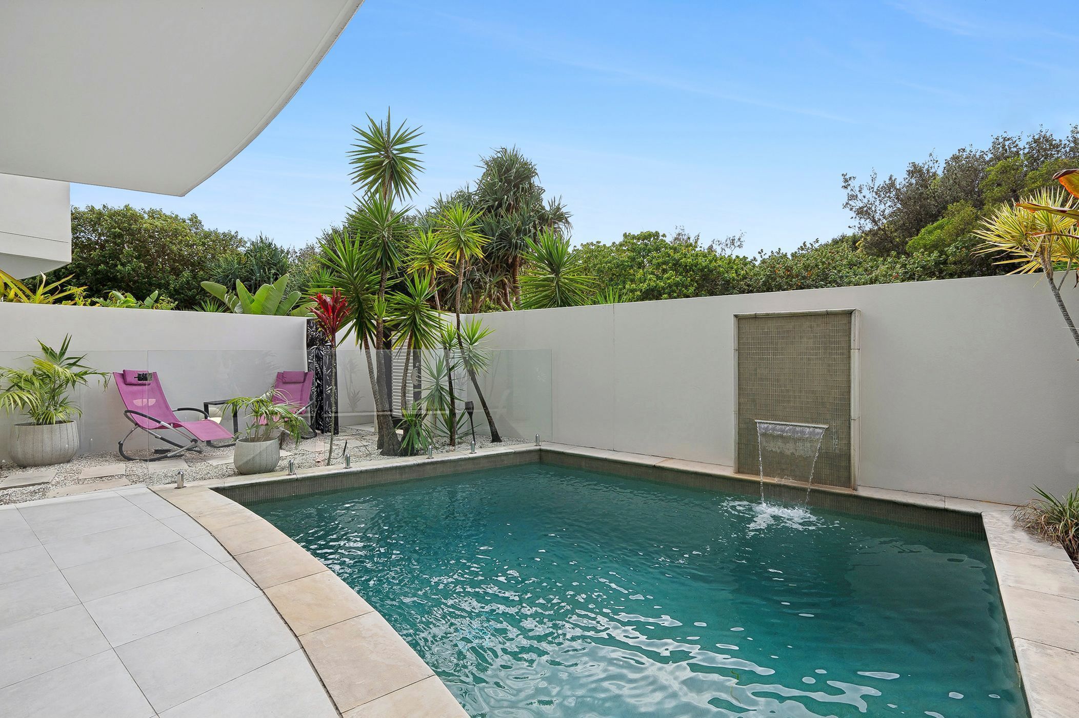 Una piscina con solárium de baldosas, rodeada de plantas en macetas y una pared blanca.