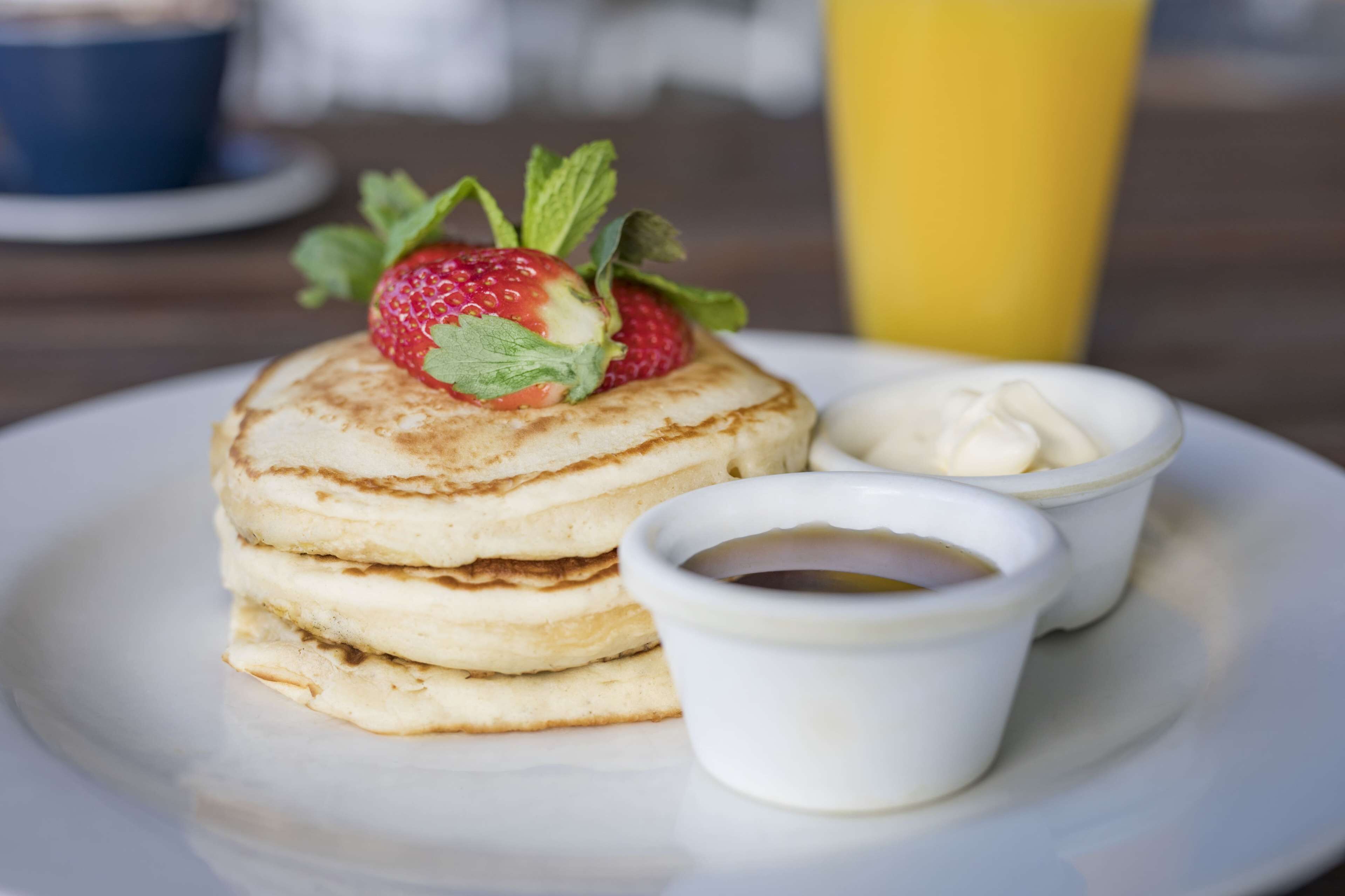 A stack of pancakes with a strawberry on top, accompanied by two small bowls of syrup and a cup of coffee.