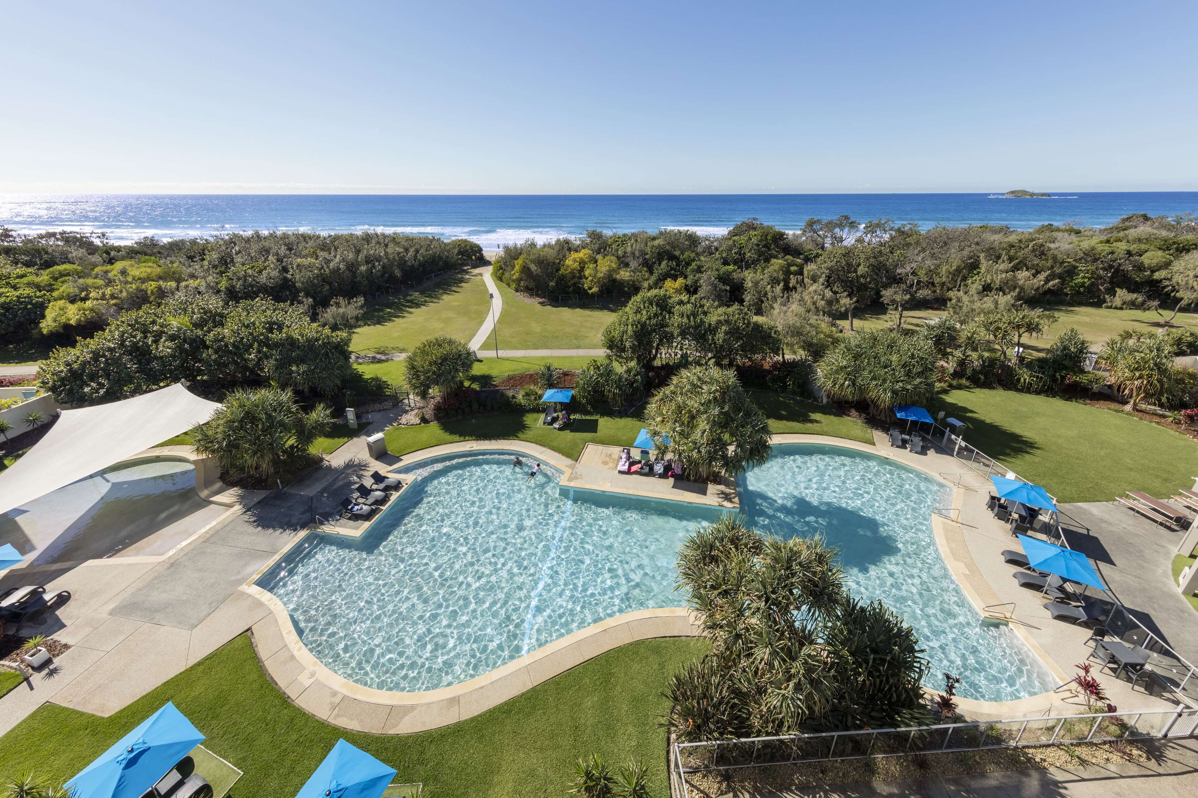 A pool area with a view of the ocean, surrounded by greenery and shaded by umbrellas.