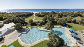 A pool area with a view of the ocean, surrounded by greenery and shaded by umbrellas.