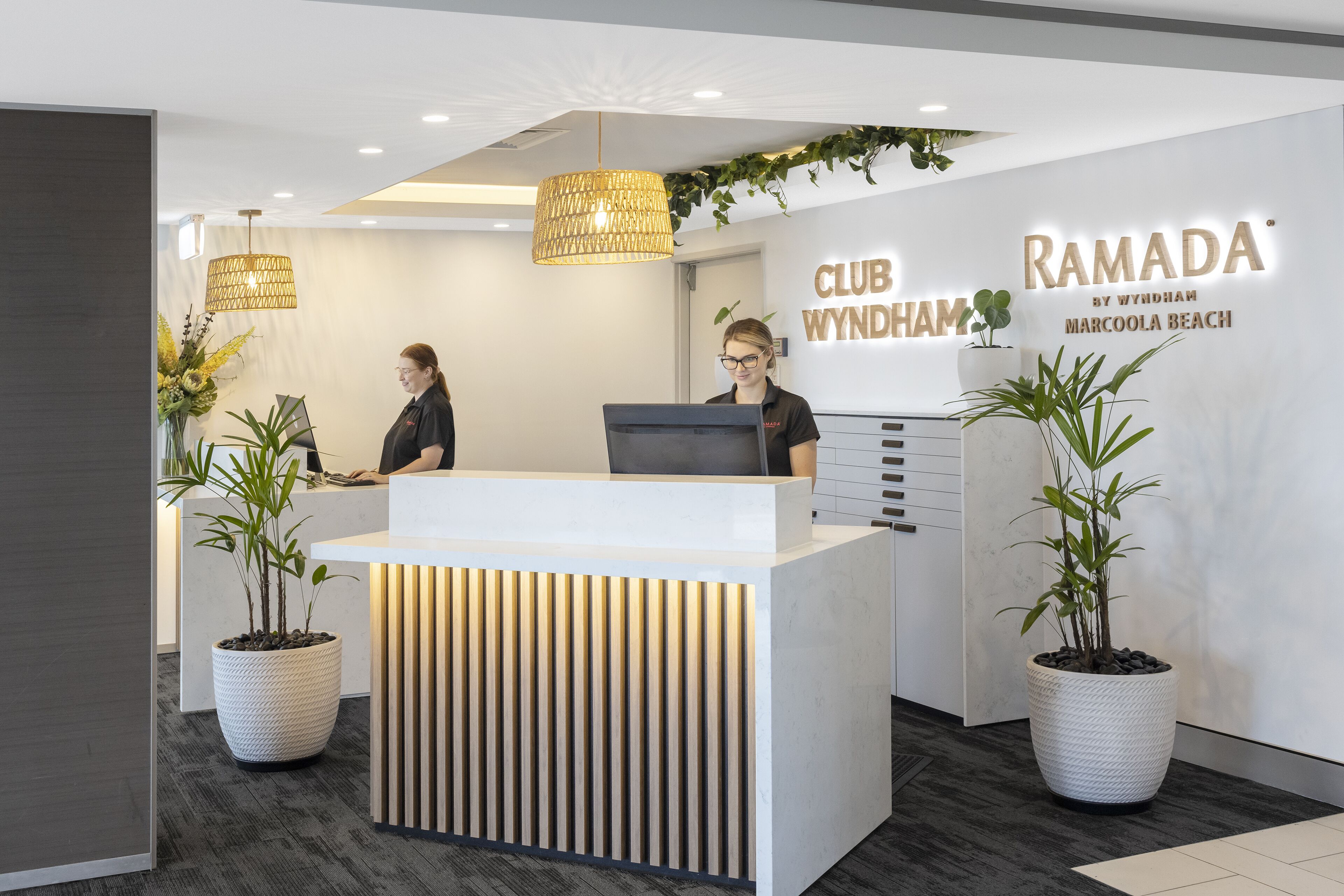 A reception desk with a modern design, featuring a white reception area, two potted plants, and a sign that reads "CLUB WYNDHAM RAMADA MARCOOLA BEACH".